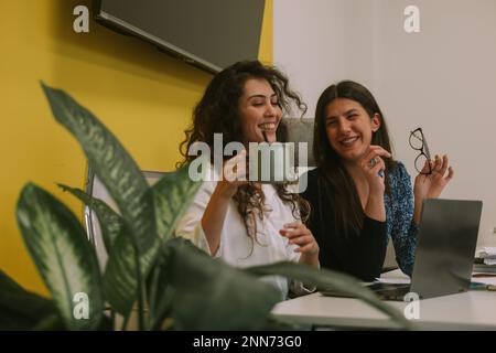 Nahaufnahme von zwei schönen Geschäftsfrauen, die lächeln und sich im Büro amüsieren, während sie sitzen Stockfoto