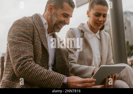 Nahaufnahme eines älteren Geschäftsmanns, der lächelt und seinem Kollegen etwas auf seinem Tablet zeigt, während er im Park sitzt Stockfoto