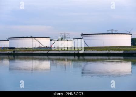 Große Rohöltanks in einem Ölterminal in der Dämmerung. Windturbinen und Hochspannungsleitungen befinden sich im Hintergrund. Stockfoto