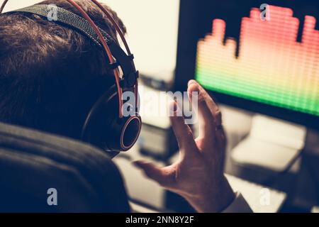 Rückansicht des Electronic Music Producers, der vor seinem Computer sitzt und sich in Kopfhörern einen neu erstellten Titel anhört. Stockfoto