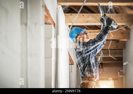 Professioneller, kaukasischer Elektriker, der an der Installation von elektrischen Verkabelungssystemen in der Decke eines neuen Wohnhauses arbeitet. Stockfoto