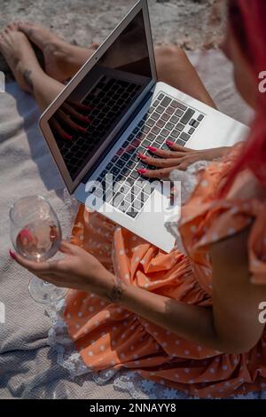 Eine junge Frau mit einem Notebook sitzt in der Natur und arbeitet Remote. Er hält ein Glas Wein in der Hand. Stockfoto