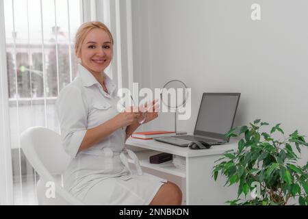 Blonde Ärztin in der Arztpraxis, lächelnd. Auf dem Tisch ist ein Computer, ein Spiegel. Das Büro hat weiße Wände, eine Blume mit grünen Blättern. Stockfoto