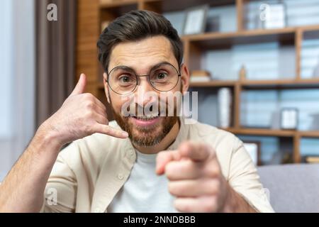 Nahaufnahme. Porträt eines jungen Mannes mit Brille, der mit der Kamera spricht. Zeigt und erklärt mit einer Call-Me-Handgeste und zeigt auf die Kamera. Stockfoto