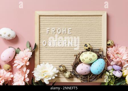 Anschreiben, Frohe Ostern - Happy Easter in deutscher Begrüßung, Ostereier und Frühlingsblumen auf pastellrosa Hintergrund. Blick von oben. Stockfoto