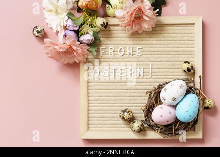 Anschreiben, Frohe Ostern - Happy Easter in deutscher Begrüßung, Ostereier und Frühlingsblumen auf pastellrosa Hintergrund. Blick von oben. Stockfoto