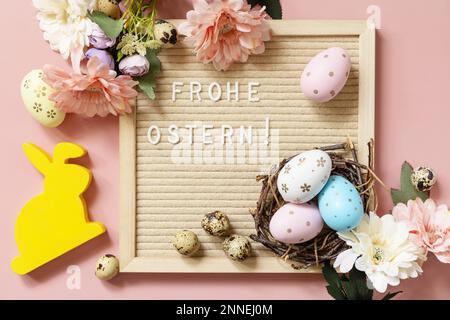 Anschreiben, Frohe Ostern - Happy Easter in deutscher Begrüßung, Ostereier und Frühlingsblumen auf pastellrosa Hintergrund. Blick von oben. Stockfoto