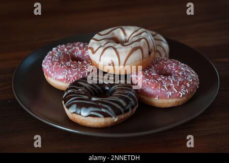 Mehrfarbige, weiße, rosafarbene Schokoladen-Donuts liegen auf einem braunen Teller. Vegetarisches Frühstück, süß. Hochwertiges Foto Stockfoto