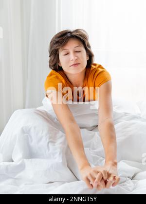 Schläfrige Frau im gelben Pyjama dehnt sich im Bett, bedeckt mit weißer Decke. Ich wache früh am Morgen auf. Die Frau schläft genug. Stockfoto