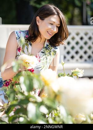 Frau bewundert blühende Rosen im öffentlichen Park. Sommerliche Stimmung. Tropische Pflanzen und Blumen blühen im Garten. Blumenzucht als Hobby. Stockfoto