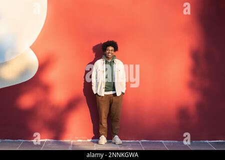 Fröhlicher, stylischer afroamerikanischer Hipster-Typ, der an der roten Wand lacht. Stockfoto