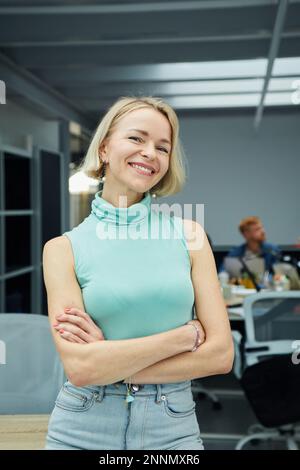 Vertikal. Lächelnde weiße Geschäftsfrau, die in die Kamera schaut und im Büro mit gekreuzten Armen steht. Stockfoto