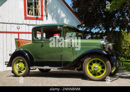 1930 Ford Model A Coupé, klassisches Auto in Grün mit schwarzen Kotflügeln und Rüttelsitz aus braunem Leder vor einer alten, weiß-roten Scheune im Sommer. Stockfoto