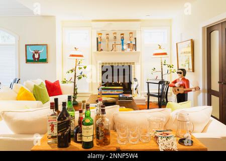 Likörtisch und weiße Polstersofas mit gelben, grünen und roten Velours-Kissen, Elvis Folk Art Skulptur, Gaskamin im Wohnzimmer. Stockfoto