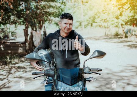 Biker auf seinem Motorrad hält Schutzhelm mit Daumen nach oben. Konzept eines lächelnden Motorradfahrers mit Schutzhelm, Biker man mit Daumen nach oben Stockfoto