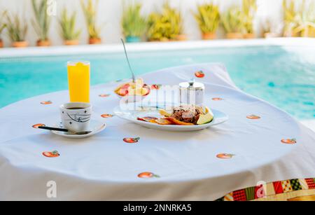 Das Frühstück wird auf dem Tisch serviert, im Hintergrund befindet sich ein Swimmingpool. Obstsalat. Das traditionelle Frühstück wird am Tisch in der Nähe des Swimmingpools serviert Stockfoto