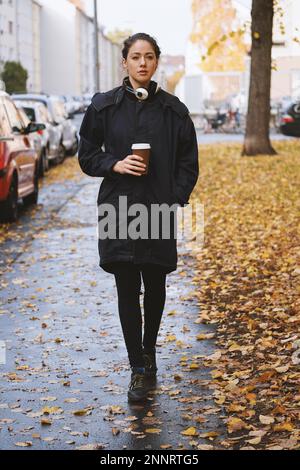 Eine junge Frau, die an einem nassen, kalten Herbsttag alleine die Straße entlang läuft, trägt einen Parka-Mantel mit Kopfhörern um den Hals und Kaffee zum Tassen in ihr Stockfoto