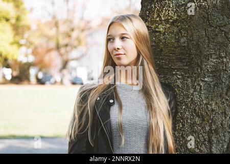 Verträumt junges Mädchen mit langen blonden Haaren und Lederjacke lehnte sich gegen den Baum an einem sonnigen Tag im Frühjahr Stockfoto
