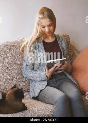 Bleiben Sie zu Hause Teenager-Mädchen Lesen Sie E-Book oder verwenden Sie einen Tablet-Computer, während Sie auf einem Sofa mit Katze sitzen - Online-Kommunikation während der Quarantäne Stockfoto