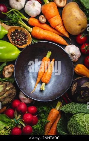 Zwei frische Bauernmöhren in klassischer Schüssel und gemischtes Bio-Gemüse auf rustikalem schwarzem Betonhintergrund. Herbsternte, vegetarische Speisen oder Stockfoto