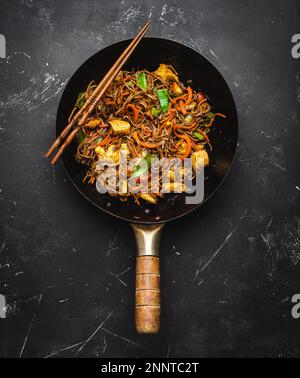 Gebratene Soba-Nudeln mit Hühnchen, Gemüse in einer alten rustikalen Wok-Pfanne, Essstäbchen auf schwarzem Steinhintergrund, Nahaufnahme, Draufsicht. Traditionell Stockfoto