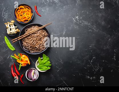 Zutaten für die Zubereitung von gebratenen Nudeln Soba. Schneiden Sie frisches Gemüse, Pilze, gekochte Soba-Nudeln in eine Schüssel mit Stäbchen, die zum Kochen bereit sind, schwarz Stockfoto