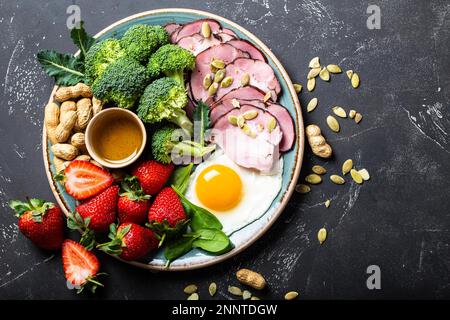 Ketogene kohlenhydratarme Diät-Konzept, Draufsicht. Platte auf steinschwarzem Hintergrund mit Keto-Lebensmitteln: Eier, Fleisch, Olivenöl, Brokkoli, Beeren, Nüsse, Samen Stockfoto