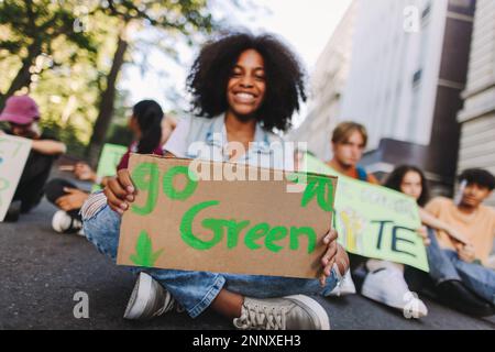 Glückliches Teenager-Mädchen, das ein „Go Green“-Poster hält, während es mit einer Gruppe von Demonstranten bei einem Protest gegen den Klimawandel sitzt. Multikulturelle Jugendaktivisten Stockfoto