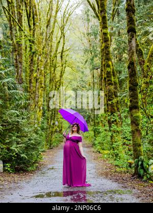 Schwangere Frau in lila Kleid mit lila Regenschirm im Wald Stockfoto