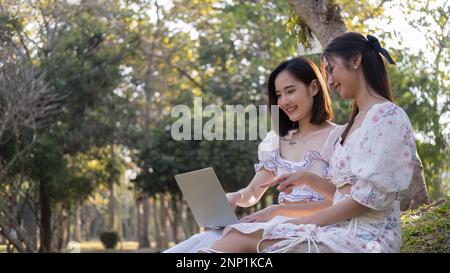 Zwei asiatische Frauen sehen sich zusammen ein Video auf einem Laptop an, während sie ein Picknick im Park genießen Stockfoto