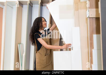 Teenager in brauner Schürze, die Sperrholz aus einem Lagerfach zieht. Atmosphäre in einer Holzmöbelfabrik. Stockfoto