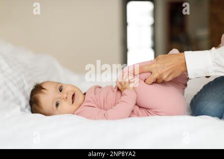 Obstipation bei Säuglingen. Fürsorgliche Mutter, die Gymnastik macht, während ein Mädchen auf dem Bett liegt, und die kleine Tochter massiert Stockfoto