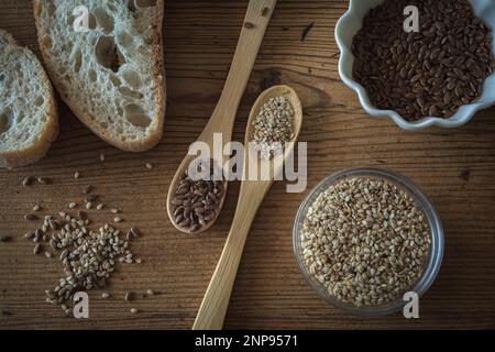 Gesundes Stillleben mit Sesam- und Leinsamen in Schüsseln und Holzlöffeln und in einer Ecke natürlichem Brot Stockfoto