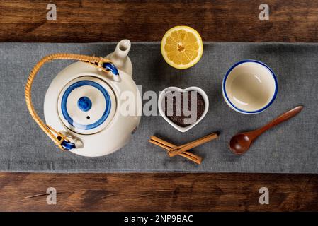 Porzellan-Teekanne neben einer kleinen herzförmigen Schüssel mit gemahlenem Tee, einer Tasse, einer halben Zitrone und Zimtstücken. Stockfoto
