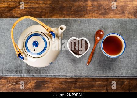 Porzellan-Teekanne neben einer kleinen herzförmigen Schüssel mit gemahlenem Tee, einem hölzernen Teelöffel und einer Tasse mit Tee. Stockfoto