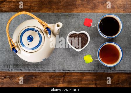 Porzellan-Teekanne neben einer kleinen herzförmigen Schüssel mit gemahlenem Tee, zwei Tassen mit verschiedenen Teesorten, dunkelrot und hellgrün. Stockfoto