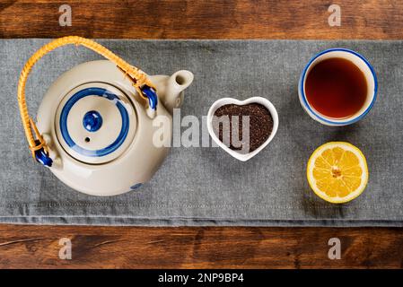 Porzellan-Teekanne neben einer kleinen herzförmigen Schüssel mit gemahlenem Tee, einer Tasse mit Tee und einer halben Zitrone. Stockfoto