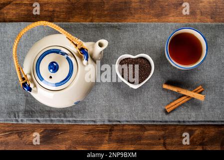 Porzellan-Teekanne neben einer kleinen herzförmigen Schüssel mit gemahlenem Tee, einer Tasse mit Tee und Zimtstangen. Stockfoto