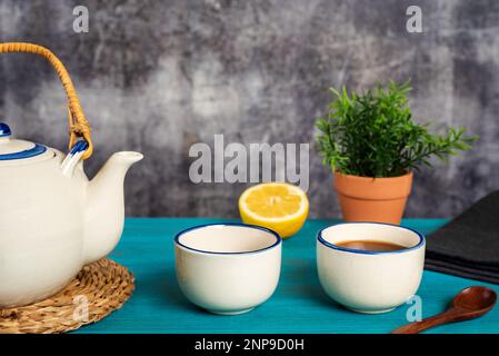 Porzellan-Teekanne neben zwei Tassen, eine mit Tee, mit einem hölzernen Teelöffel und einer halben Zitrone, auf einem blauen Holztisch. Stockfoto