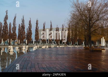 Russland, Krasnodar - 04. Januar 2023: Eine hölzerne braune Plattform in der Nähe eines künstlichen Teiches mit blumenförmigen Laternen und Pyramideneichen um den Perime Stockfoto