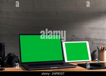 Chroma Key grüner Bildschirm, angewinkelter Laptop und digitales Tablet auf Tisch mit digitaler Fotoausrüstung auf Holztisch vor Betonwand. Stockfoto