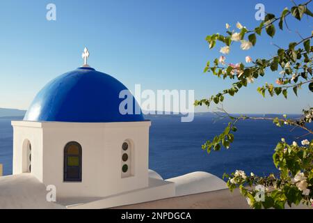 Ein Blick auf eine traditionelle blaue Kuppelkirche bei Sonnenschein am frühen Morgen mit der Caldera und dem Meer dahinter. Stockfoto