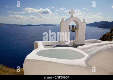 Ein Blick auf einen Glockenturm hoch oben auf dem Dach einer traditionellen griechisch-orthodoxen Kirche mit der Caldera und dem Meer dahinter. Stockfoto