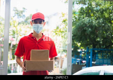 Asiatischer Liefermann trägt Gesichtsmaske in roter Uniform liefern Service Paketbox an den Kunden zu Hause Stockfoto