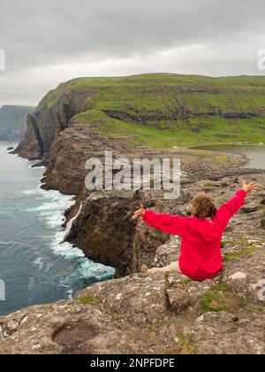 Ein Tourist in roter Jacke sitzt an einem felsigen Ufer über dem Bøsdalafossur Wasserfall, der vom See Sørvágsvatn / Leitisvatn in den Atlantik mündet Stockfoto