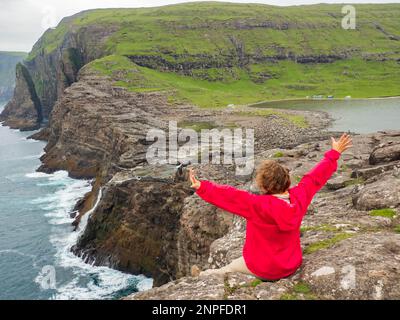 Ein Tourist in roter Jacke sitzt an einem felsigen Ufer über dem Bøsdalafossur Wasserfall, der vom See Sørvágsvatn / Leitisvatn in den Atlantik mündet Stockfoto