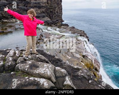 Ein Tourist in roter Jacke sitzt an einem felsigen Ufer über dem Bøsdalafossur Wasserfall, der vom See Sørvágsvatn / Leitisvatn in den Atlantik mündet Stockfoto