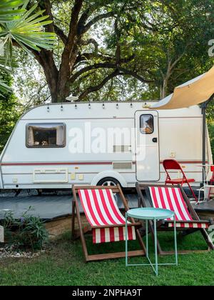 Leere Liegestühle am Strand und Klappstühle mit Tisch vor dem Wohnwagenparkplatz im Garten, vertikaler Stil. Entspannung, Camping und Schlafen in der Mo Stockfoto