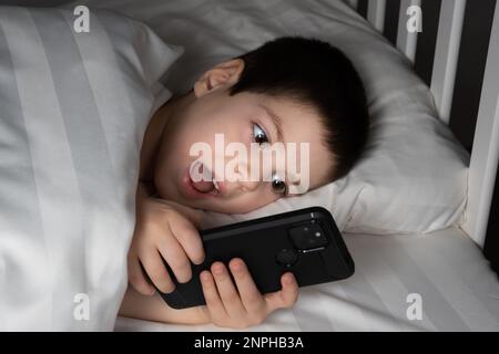 Ein 5-jähriger Junge spielt Spiele auf seinem Handy, während er im Bett liegt. Geräte im Bett vor dem Schlafengehen bei einem Kind Stockfoto