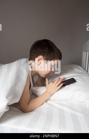 Ein 5-jähriger Junge spielt Spiele auf seinem Handy, während er im Bett liegt. Geräte im Bett vor dem Schlafengehen bei einem Kind Stockfoto